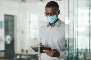 Koruyucu yüz maskesi takan genç Afrikalı işadamı ofiste dururken dijital tablet üzerinde çalışıyor.