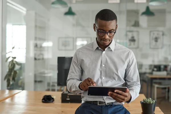 在一个现代化的大办公室里 年轻的非洲商人靠着桌子 用数字平板电脑在网上工作 — 图库照片