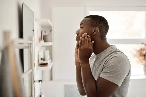 朝バスルームミラーの前に立っている間に彼の顔に触れる若いアフリカ人男性 — ストック写真