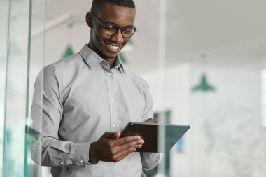 Gülümseyen genç Afrikalı işadamı modern bir ofiste tek başına duruyor ve dijital tabletle internette çalışıyor.