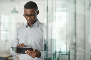 Genç Afrikalı işadamı modern bir ofiste tek başına duruyor ve dijital tabletle internette çalışıyor.