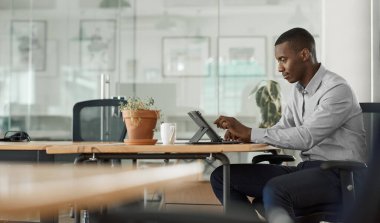 Genç Afrikalı işadamı modern bir ofiste masada oturuyor ve dijital tabletle internette çalışıyor.