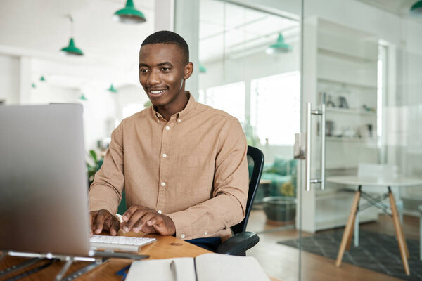 Smiling young African businessman sitting at his desk in a modern office and working on a computer