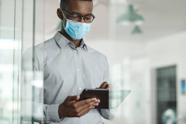 Genç Afrikalı iş adamı koruyucu bir maske takıyor ve modern bir ofiste çalışırken tablet üzerinde çalışıyor.