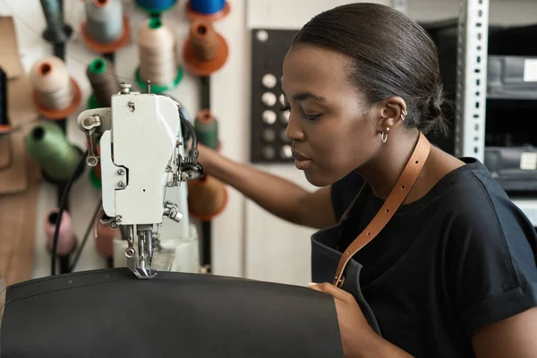 Young African female artisan sewing pieces of leather while using a sewing machine in her workshop