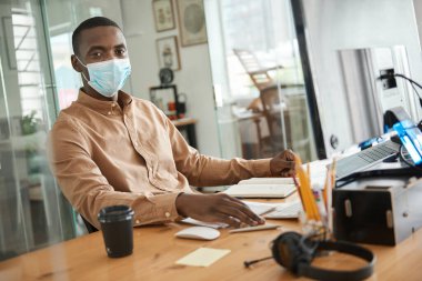 Ofisteki masasında otururken koruyucu maske takan genç bir Afrikalı işadamının portresi.