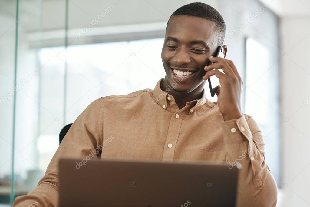 Young African businessman talking on a cellphone and working on a laptop while sitting at his office desk