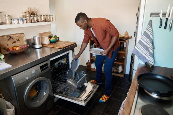 非洲年轻人在家里的厨房里把碗碟堆在洗碗机里做家务活 — 图库照片