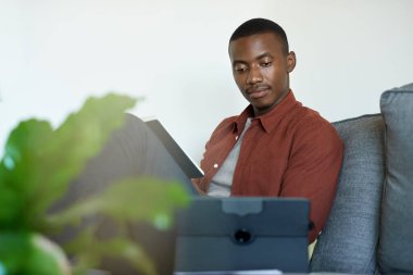Genç Afrikalı adam evinden oturma odasındaki kanepesinde çalışırken iki dijital tablet kullanıyor.