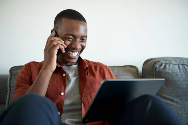 Jovem Africano Rindo Conversando Seu Telefone Usando Tablet Digital Enquanto — Fotografia de Stock