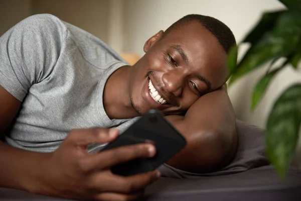 Uśmiechnięty Młody Afrykanin Czytający Sms Komórce Leżąc Rano Swoim Łóżku — Zdjęcie stockowe