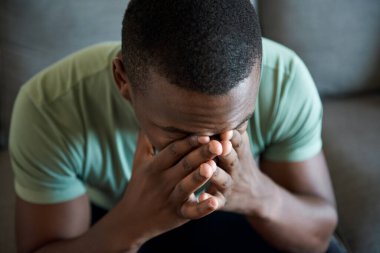 Kafası karışmış genç Afrikalı bir adam oturma odasındaki kanepede tek başına oturuyor.