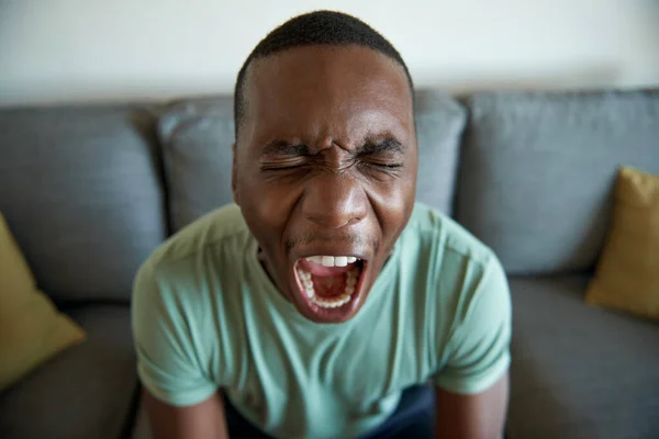 一个年轻的非洲男人坐在客厅沙发上痛苦地尖叫着的衣服 — 图库照片