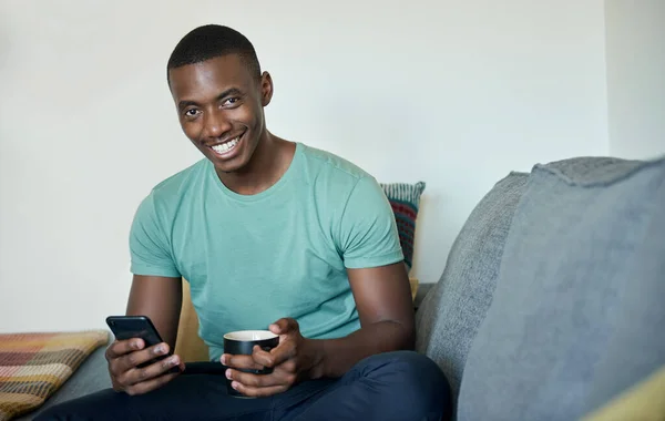 一个微笑的非洲年轻人坐在家里沙发上 用手机发短信 喝咖啡的画像 — 图库照片