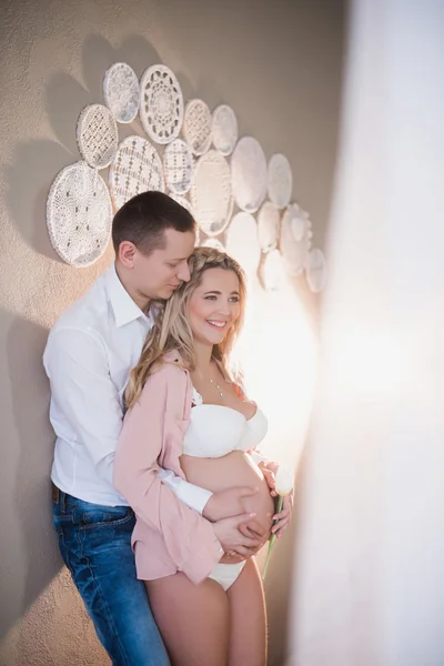 Esposo abrazando a su esposa embarazada en lencería blanca sobre un fondo claro —  Fotos de Stock