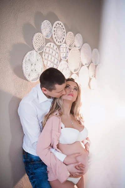 Esposo abrazando a su esposa embarazada en lencería blanca sobre un fondo claro — Foto de Stock