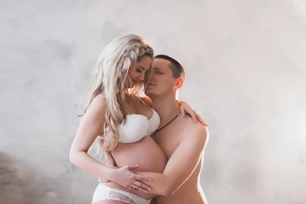 Esposo abrazando a su esposa embarazada en lencería blanca sobre un fondo claro — Foto de Stock