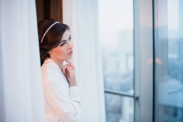 Retrato de una hermosa joven con una túnica blanca en el dormitorio cerca de la ventana —  Fotos de Stock