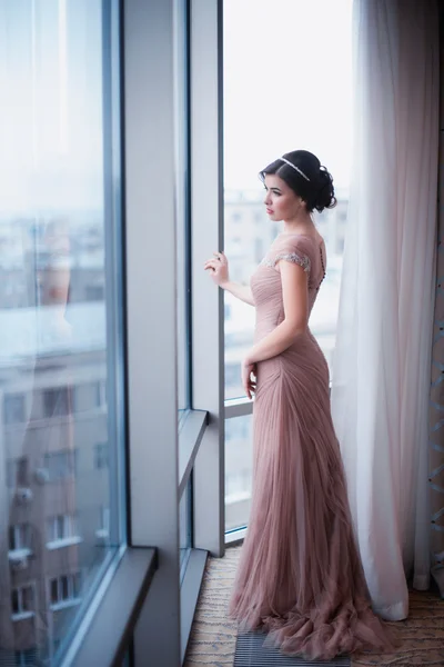 Женщина в красивом платье розового цвета стоит у окна — стоковое фото