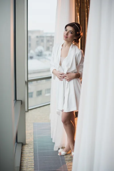 Portrait of a beautiful young girl in a white robe in the bedroom near the window
