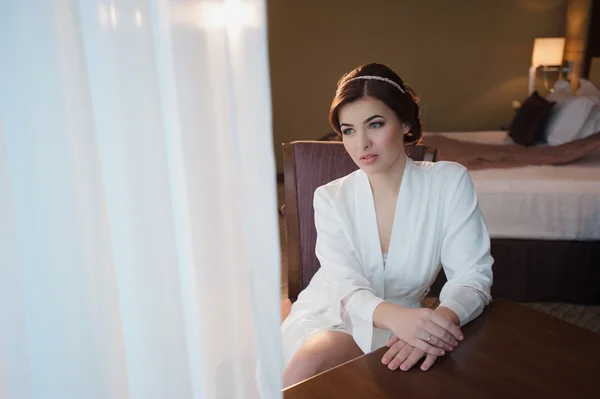 Retrato de una hermosa joven con una túnica blanca en el dormitorio cerca de la ventana —  Fotos de Stock