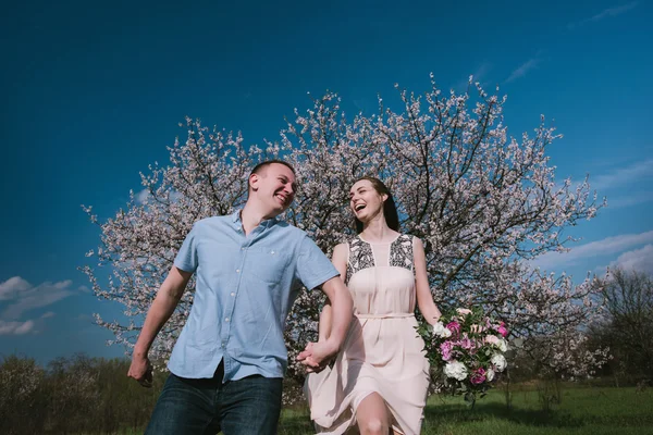Счастливая пара, бегущая в цветущем саду, держась за руки на голубом фоне неба — стоковое фото