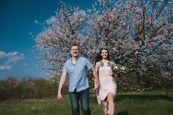Счастливая пара, бегущая в цветущем саду, держась за руки на голубом фоне неба — стоковое фото