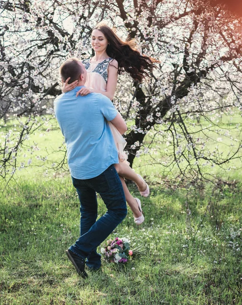 Мужчина, держащий счастливую женщину на руках в цветущем весеннем саду — стоковое фото