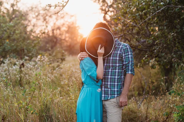 The guy and the girl standing on nature, embrace and kiss under the guise of a wide hat — Stock Photo, Image