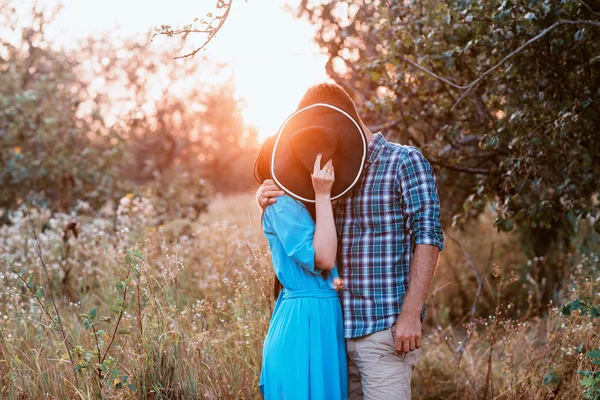 El chico y la chica de pie en la naturaleza, abrazar y besar bajo la apariencia de un sombrero ancho — Foto de Stock