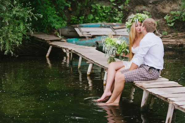 Young couple in a wreath with a bouquet on a wooden bridge laughing — Stock Photo, Image