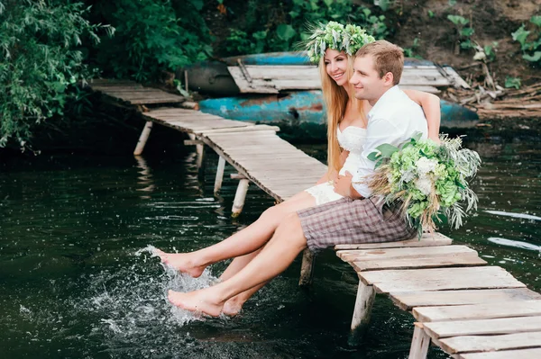 Casal jovem em uma grinalda com um buquê em uma ponte de madeira rindo — Fotografia de Stock