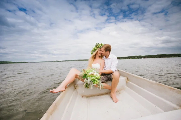 Glada unga par med en bukett och en krans kram sitter i en båt på sjön — Stockfoto