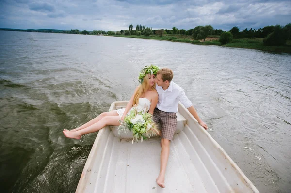 Glada unga par med en bukett och en krans kram sitter i en båt på sjön — Stockfoto