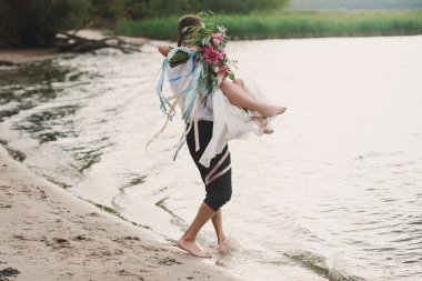 Genç yakışıklı damat gelini güzel çiçekleri büyük bir buket ile sahilde onun kollarında tutar