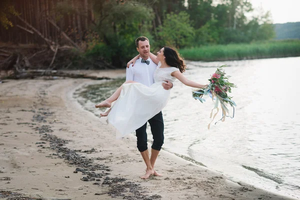 年轻英俊的新郎抱他的新娘在怀里是在沙滩上一大束美丽的鲜花 — 图库照片