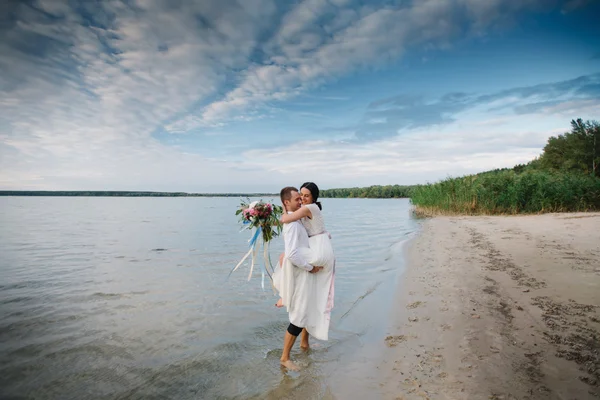 Junger hübscher Bräutigam hält seine Braut am Strand mit einem großen Strauß schöner Blumen in den Armen — Stockfoto