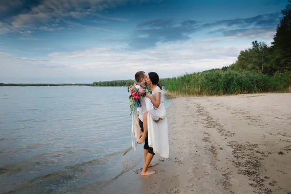 Junger hübscher Bräutigam hält seine Braut am Strand mit einem großen Strauß schöner Blumen in den Armen — Stockfoto