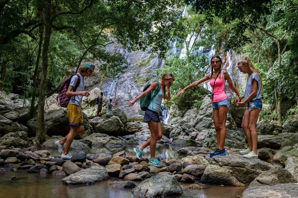 Young tourists are moving across the creek on the rocks in the jungle holding hands of each other