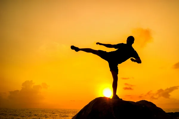 Silueta de un hombre haciendo ejercicios sobre rocas junto al mar al amanecer —  Fotos de Stock