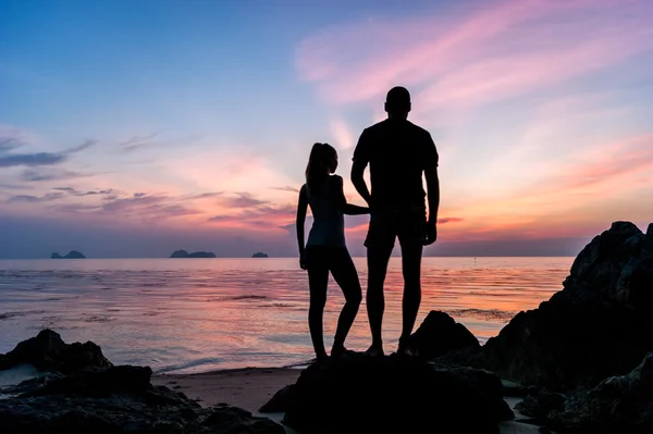 Силуэт молодой пары, стоящей на пляже, держась за руки и наблюдая за тропическим закатом — стоковое фото