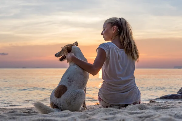 年轻女人与狗坐在沙滩上，看日落 — 图库照片
