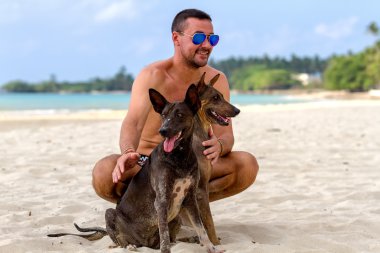 Adam deniz arka plan üzerinde iki köpek ile tropikal bir kumsalda oturur