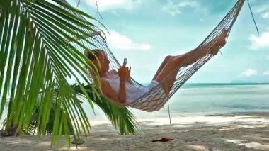 Telefonu olan genç bir kadın tropik bir plajda bir hamakta yatıyor.