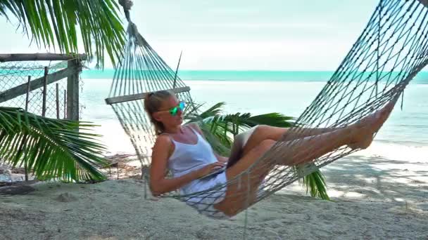 Молода жінка, що працює на ноутбуці, лежить в гамаку на тропічному пляжі — стокове відео