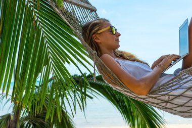Tropikal bir plajda bir hamakta dizüstü bilgisayarda çalışan genç kadın yatıyor