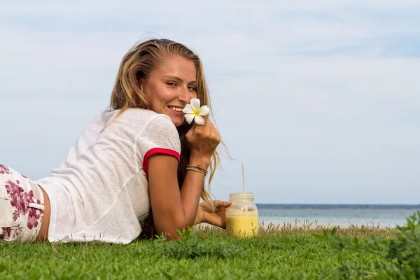 Jovem mulher com um copo de smoothie está descansando na grama em um fundo de uma praia tropical e mar em Koh Samui, Tailândia — Fotografia de Stock