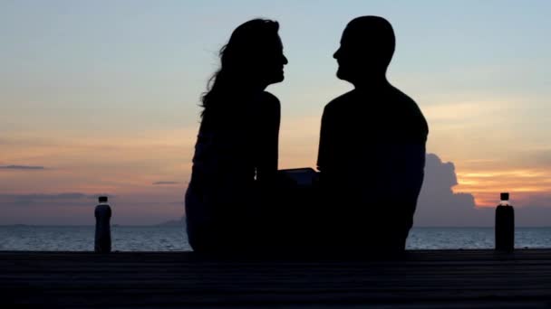 Молода пара сидить на пірсі на фоні заходу сонця — стокове відео
