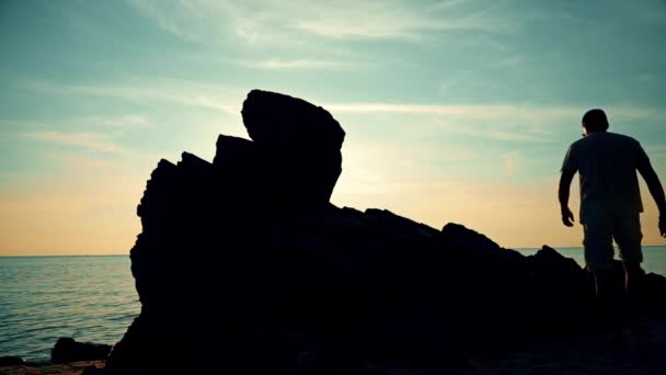 男がビーチで夕暮れ時岩に登る — ストック動画