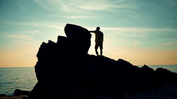 El hombre sube a una roca al atardecer en la playa — Vídeos de Stock